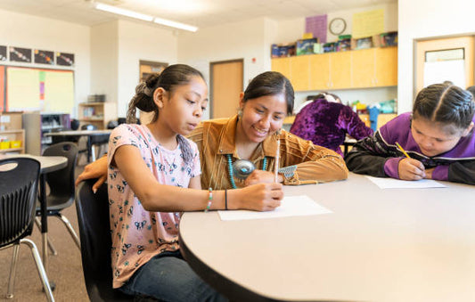 Three students sit at a round table in a classroom, working on papers with focused expressions. Shelves with books and colorful signs are in the background.