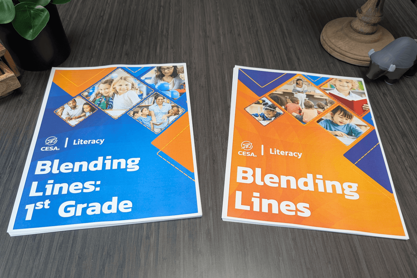 Two printed educational booklets from CESA 6 are displayed on a dark wood table. The booklet on the left has a blue cover with the title “Blending Lines: 1st Grade” and features photos of educators and children in learning settings. The booklet on the right has an orange cover titled “Blending Lines” and includes similar photo elements. The Buffalo Stress Reliever is visible in the upper-right corner next to a wooden globe base.