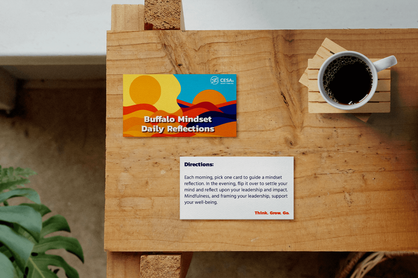 A wooden tabletop displays two rectangular cards and a white coffee mug on a wooden coaster. The top card has a bold, colorful design with abstract sun and mountain shapes and reads “Buffalo Mindset Daily Reflections” along with the CESA 6 logo. The bottom card provides directions in black text. In the lower right corner of the card, it reads: Think. Grow. Go. in red text. A green houseplant leaf enters the frame from the bottom left corner.