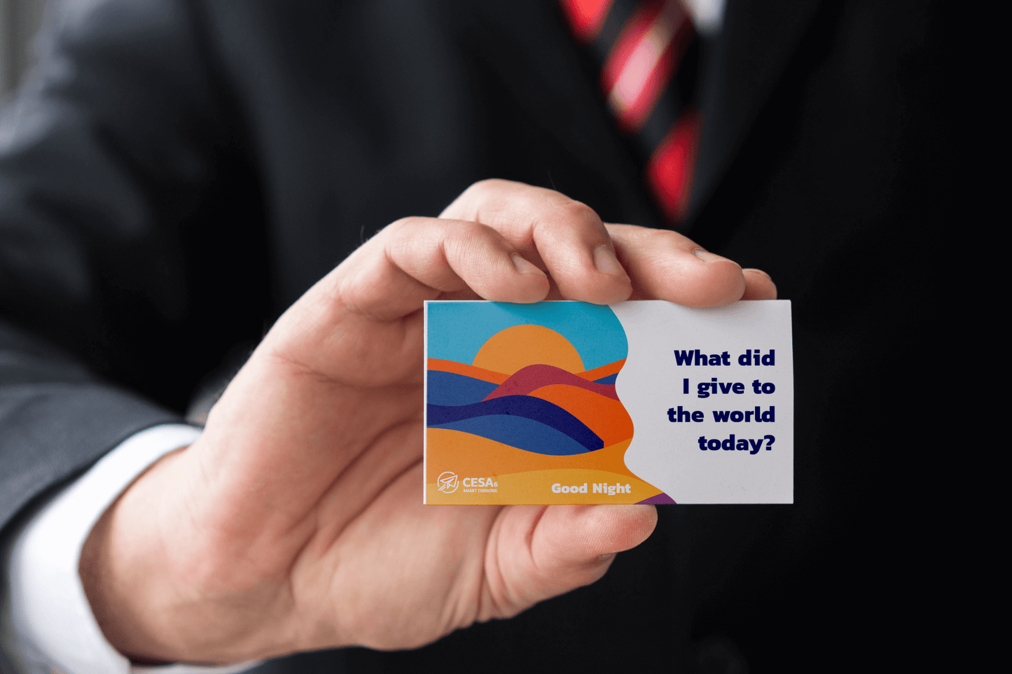 A hand in a dark suit with a red-striped tie holds a small rectangular card in focus. The card features a colorful abstract landscape with layered hills and a sun in warm tones. The right side of the card contains bold blue text that reads:
"What did I give to the world today?"
In the bottom left corner is the CESA 6 logo, and next to it in yellow text is the phrase "Good Night." The background is softly blurred to emphasize the card.