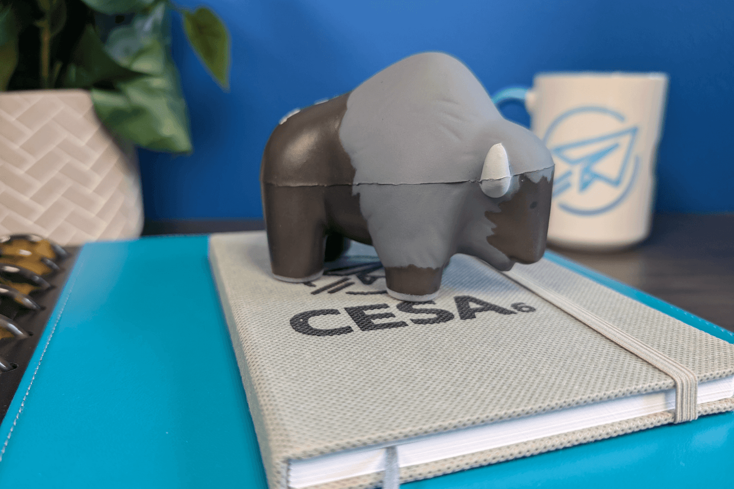 A small buffalo-shaped stress reliever toy is positioned on top of a textured notebook that has the CESA 6 logo printed on the cover. The scene is set on a desk with a bright blue folder beneath the notebook. In the background, a white ceramic mug with a blue paper airplane logo and a leafy plant in a white woven pot are visible. The overall setting suggests a workspace or office environment.
