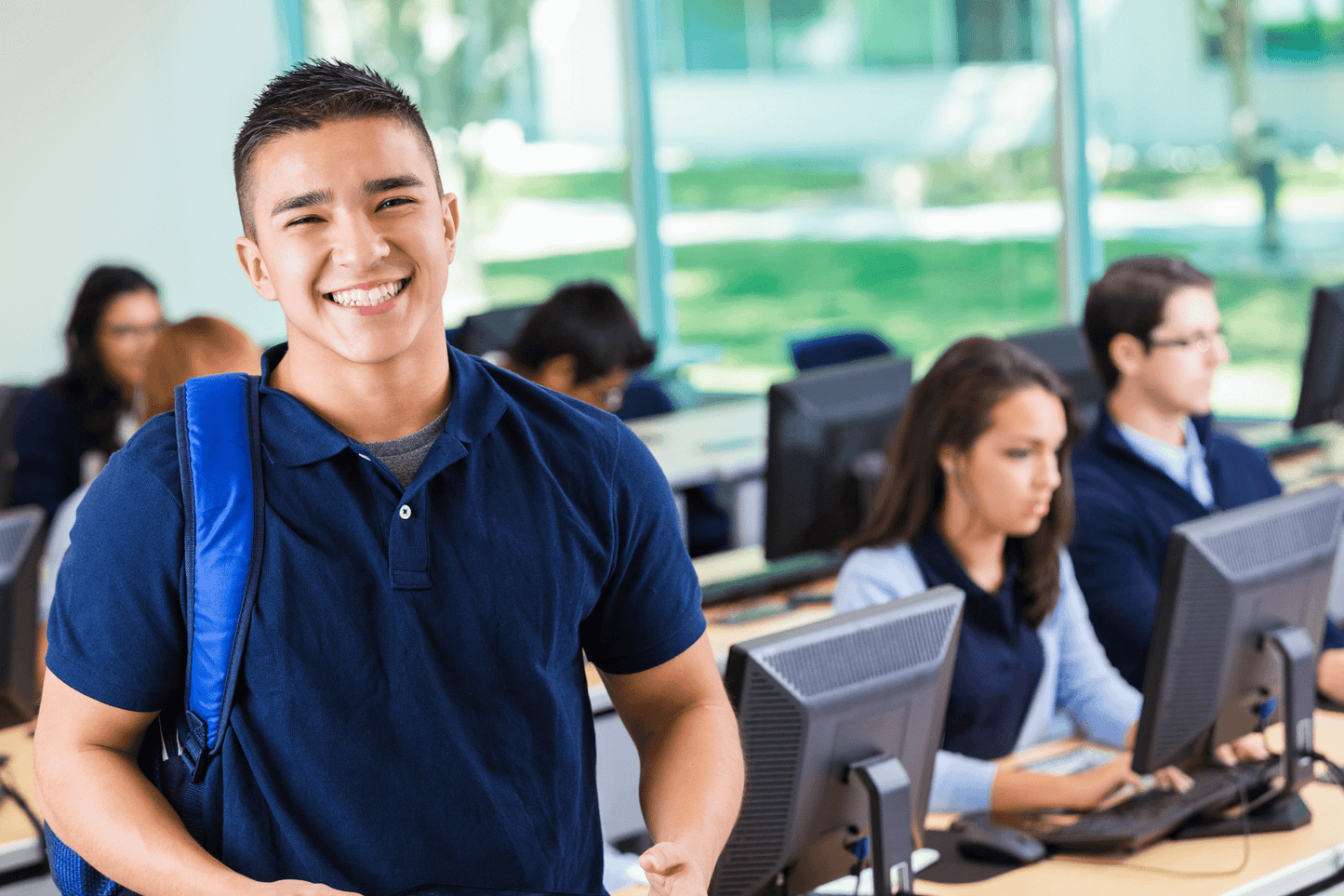 A smiling student wearing a dark blue shirt and backpack stands in a computer lab, looking at the camera. In the background, other students are seated at desktop computers, focused on their screens. The room is brightly lit with large windows showing greenery outside.