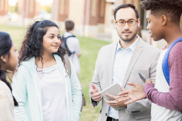 A teacher discusses with three students outside of a school building, holding a tablet while the students engage in conversation.