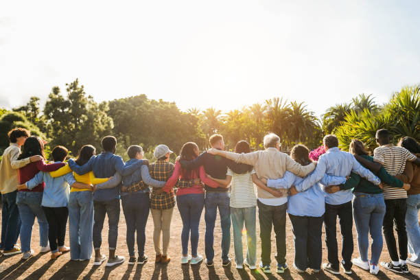 A diverse group of people stands together, arm in arm, facing away from the camera, with sunlight shining in the background.