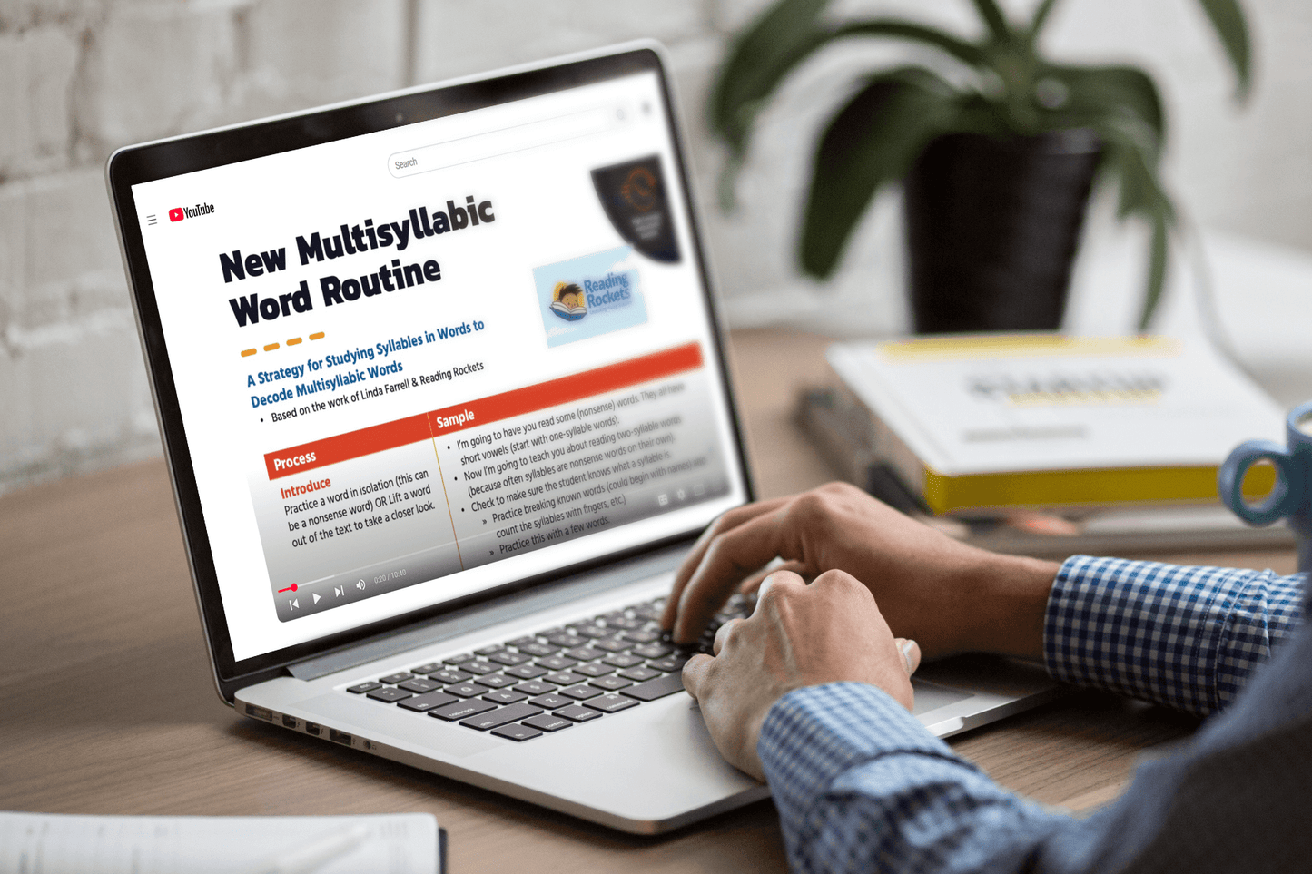 A person in a checkered shirt watches a YouTube video titled "New Multisyllabic Word Routine" on a laptop, with books and a plant nearby.