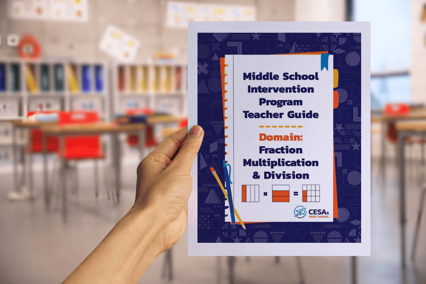 A hand holds a booklet titled “Middle School Intervention Program Teacher Guide – Domain: Fraction Multiplication & Division.” The cover is designed with a blue patterned border, orange and blue accents, and small graphics of fraction bars and grids representing multiplication of fractions. The CESA 6 logo appears at the bottom. The background shows a blurred classroom with red chairs and colorful storage bins.