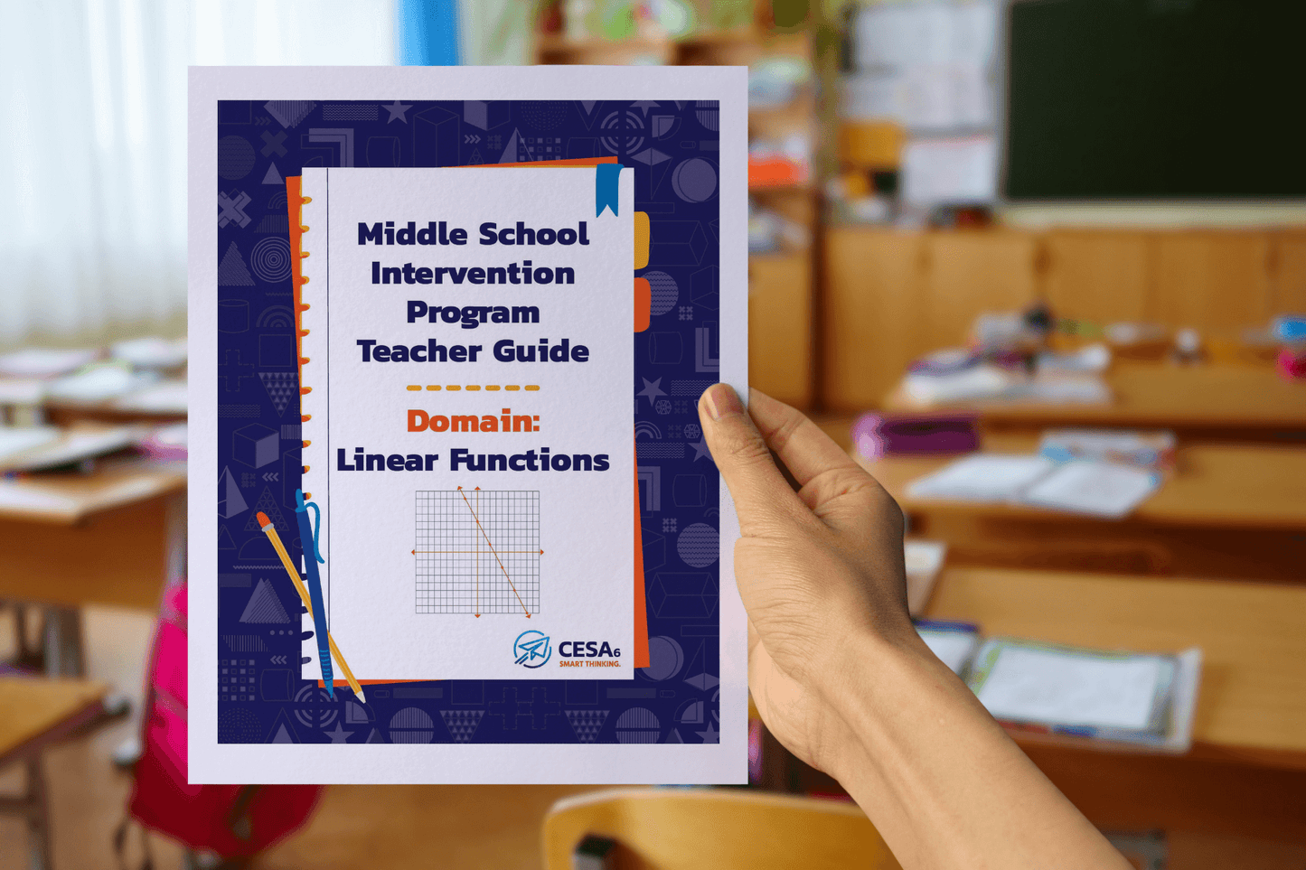 A person holds a booklet titled “Middle School Intervention Program Teacher Guide – Domain: Linear Functions”. The cover is decorated with classroom-themed graphics and features a coordinate grid with a downward-sloping line, representing a linear function. The background shows a classroom with wooden desks, papers, and school supplies.