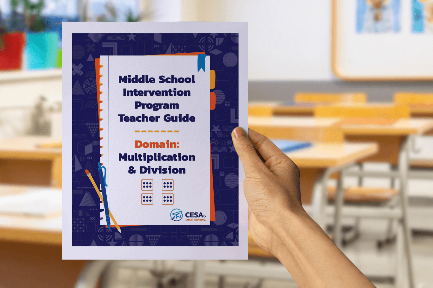 A person is holding a booklet titled "Middle School Intervention Program Teacher Guide – Domain: Multiplication & Division" in a classroom setting. The cover features a dark blue patterned border with orange and blue accents and shows small dice illustrations under the title. The blurred background displays desks, chairs, and classroom decorations.