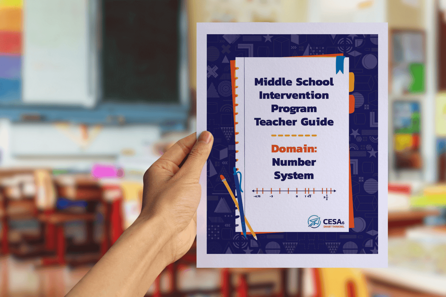 A person is holding up a booklet titled "Middle School Intervention Program Teacher Guide – Domain: Number System" in a classroom setting. The cover is designed with a dark blue patterned border, orange and blue accents, and features a number line graphic at the bottom. The blurred background shows colorful classroom decorations, bookshelves, and desks.