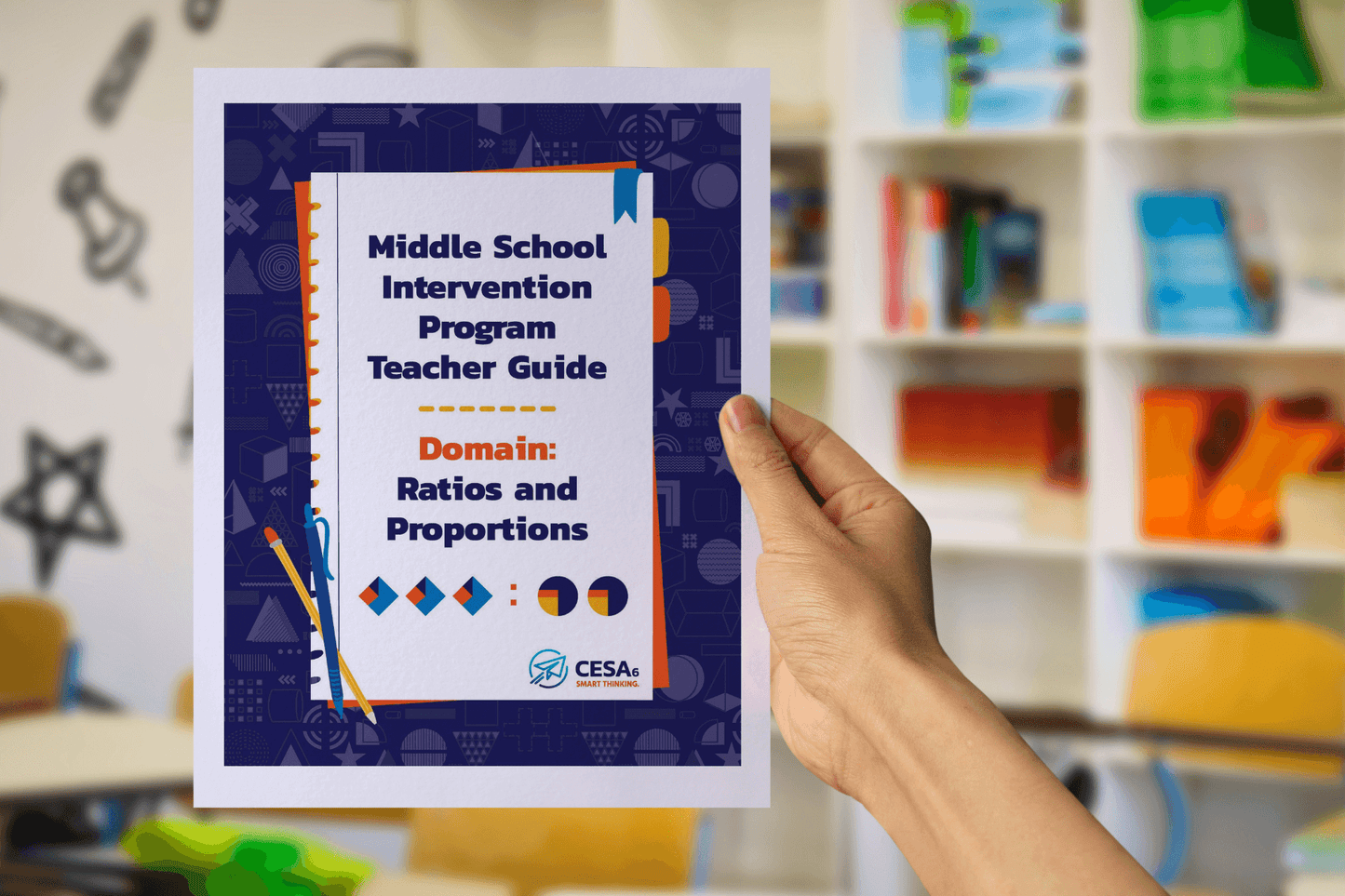 A hand holds up a booklet titled Middle School Intervention Program Teacher Guide: Domain – Ratios and Proportions. The cover features colorful math-themed graphics, including geometric shapes and ratio symbols. In the background, blurred bookshelves and classroom chairs are visible, suggesting a school setting.