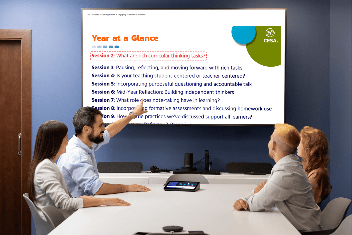 Four colleagues sit at a conference table, attentively viewing a large screen displaying a professional learning slide titled "Year at a Glance." One participant gestures toward the screen, highlighting a list of CESA 6 training sessions such as “What are rich curricular thinking tasks?” and “What role does note-taking have in learning?”