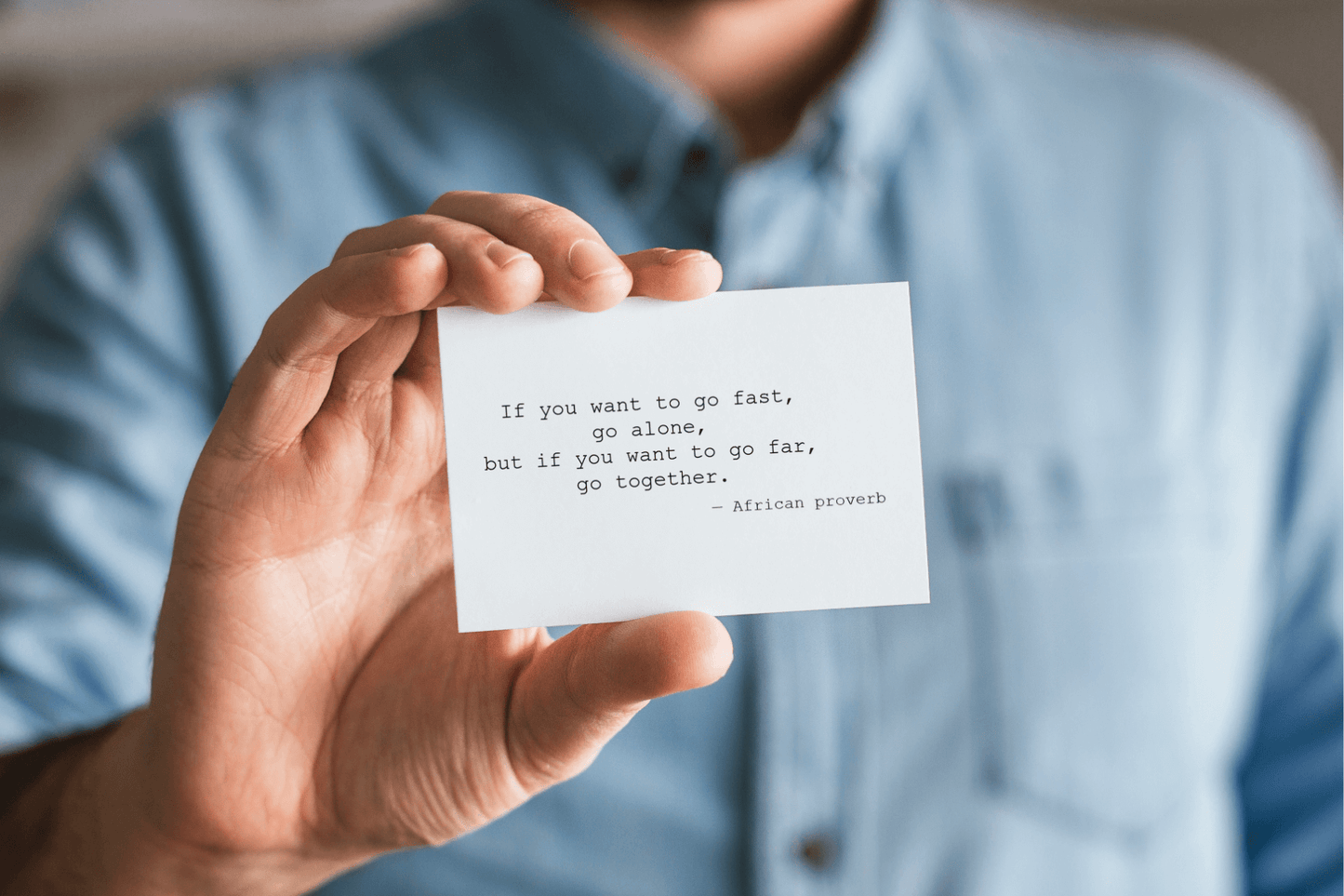 A person in a light blue shirt is holding a small white card close to the camera. The card displays a quote in typewriter-style font: “If you want to go fast, go alone, but if you want to go far, go together. — African proverb.”