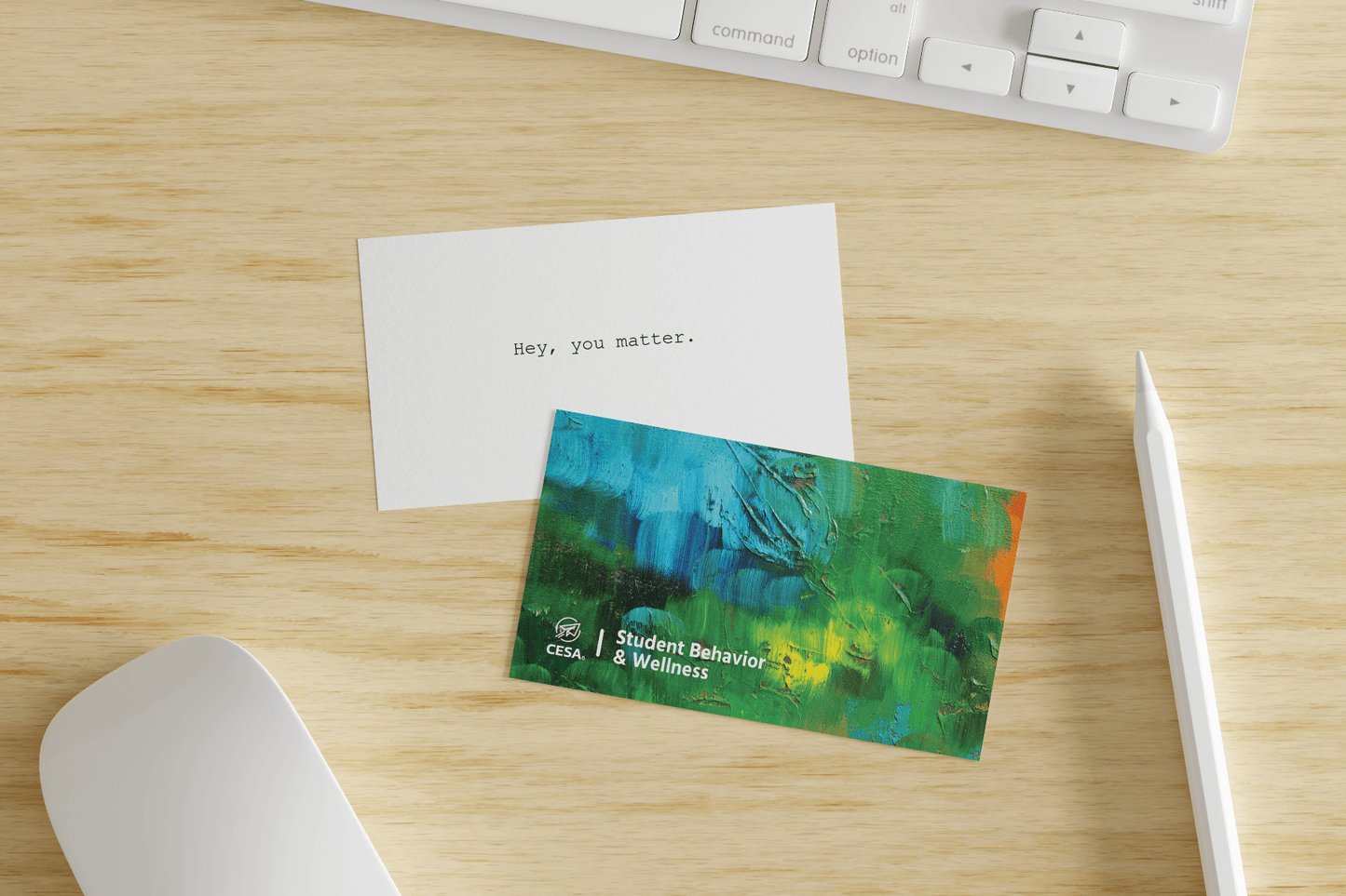 Two cards rest on a wooden desk near a white keyboard, stylus, and mouse. One card has a plain white background with the typed message: “Hey, you matter.” The second card features a textured, abstract painting in blue and green tones and is labeled “CESA 6 | Student Behavior & Wellness.”