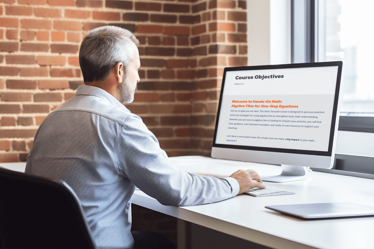 A man with gray hair sits at a white desk, working on a computer displaying "Course Objectives." The setting is modern, with a brick wall backdrop.