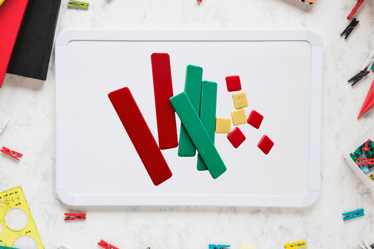 Colorful geometric shapes, including red and green rectangles and small red and yellow squares, are arranged on a whiteboard. The background has scattered stationery items, creating a playful and creative atmosphere.