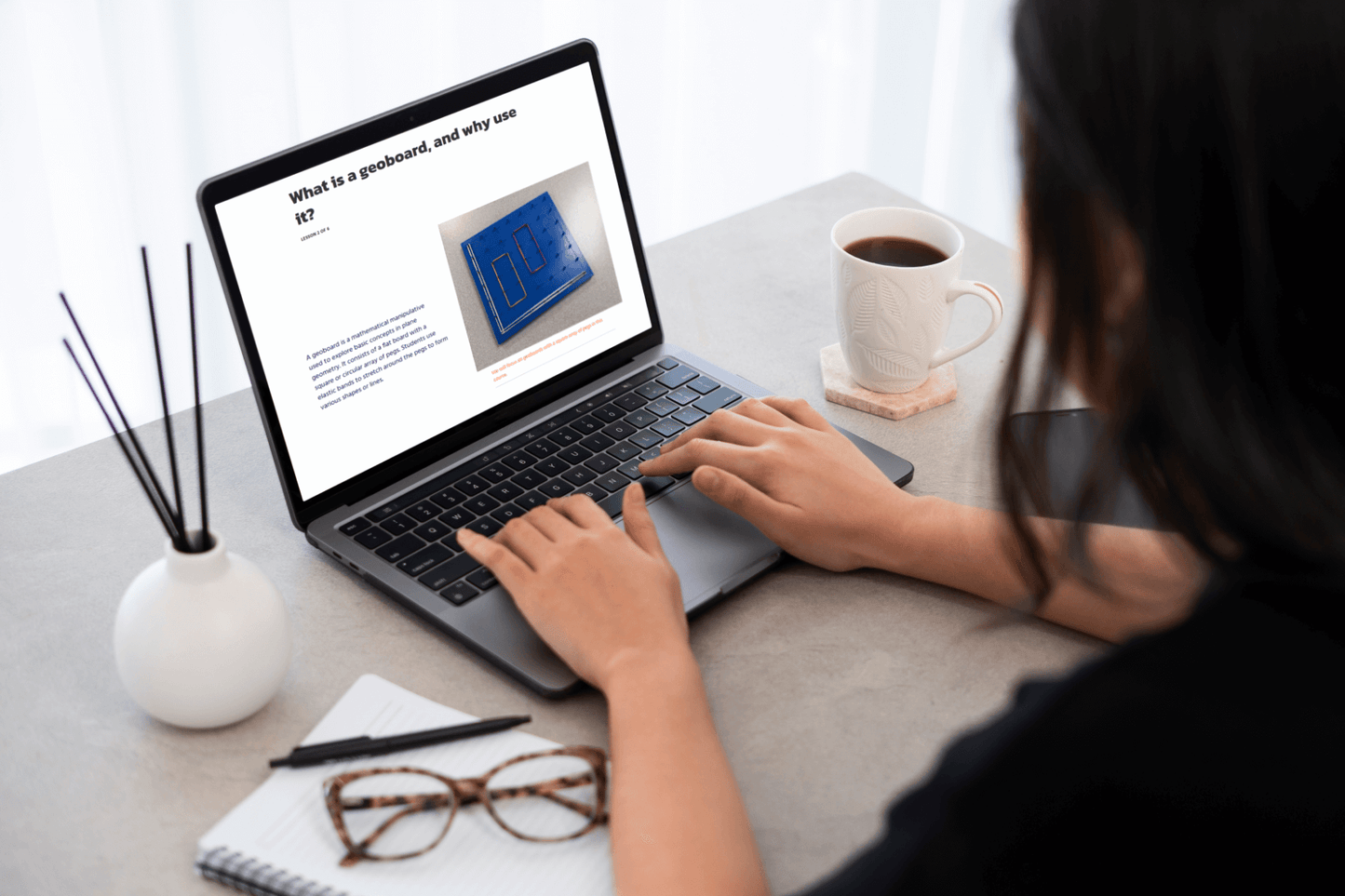 A person working on a laptop at a desk, reading an article about geoboards. Nearby are a notebook, glasses, and a cup of coffee, creating a focused workspace.