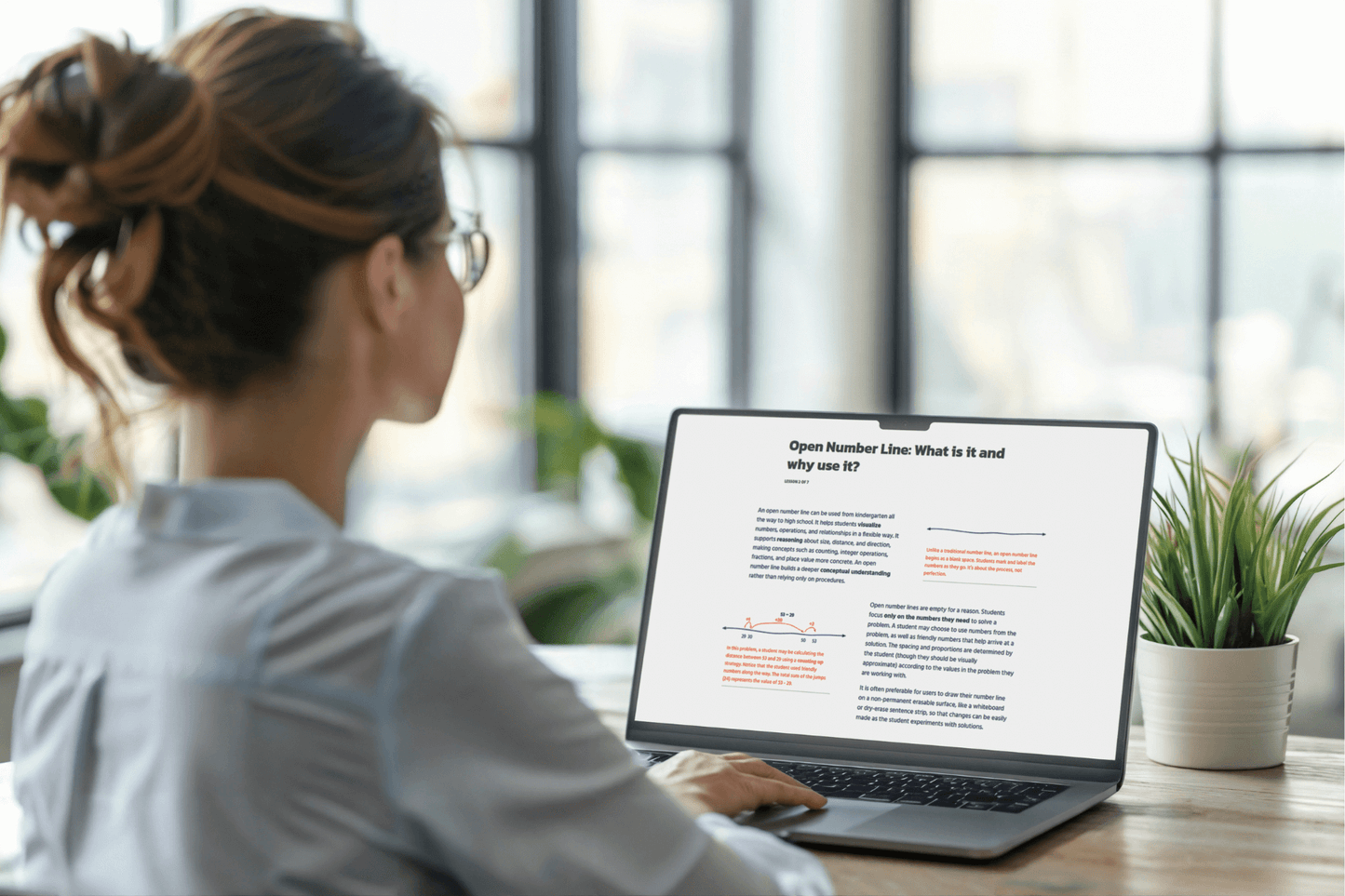 A woman with glasses sits at a wooden table, focused on a laptop screen displaying a document titled "Open Number Line: What is it and why use it?" Two small plants nearby add a touch of green. The scene is calm and studious.