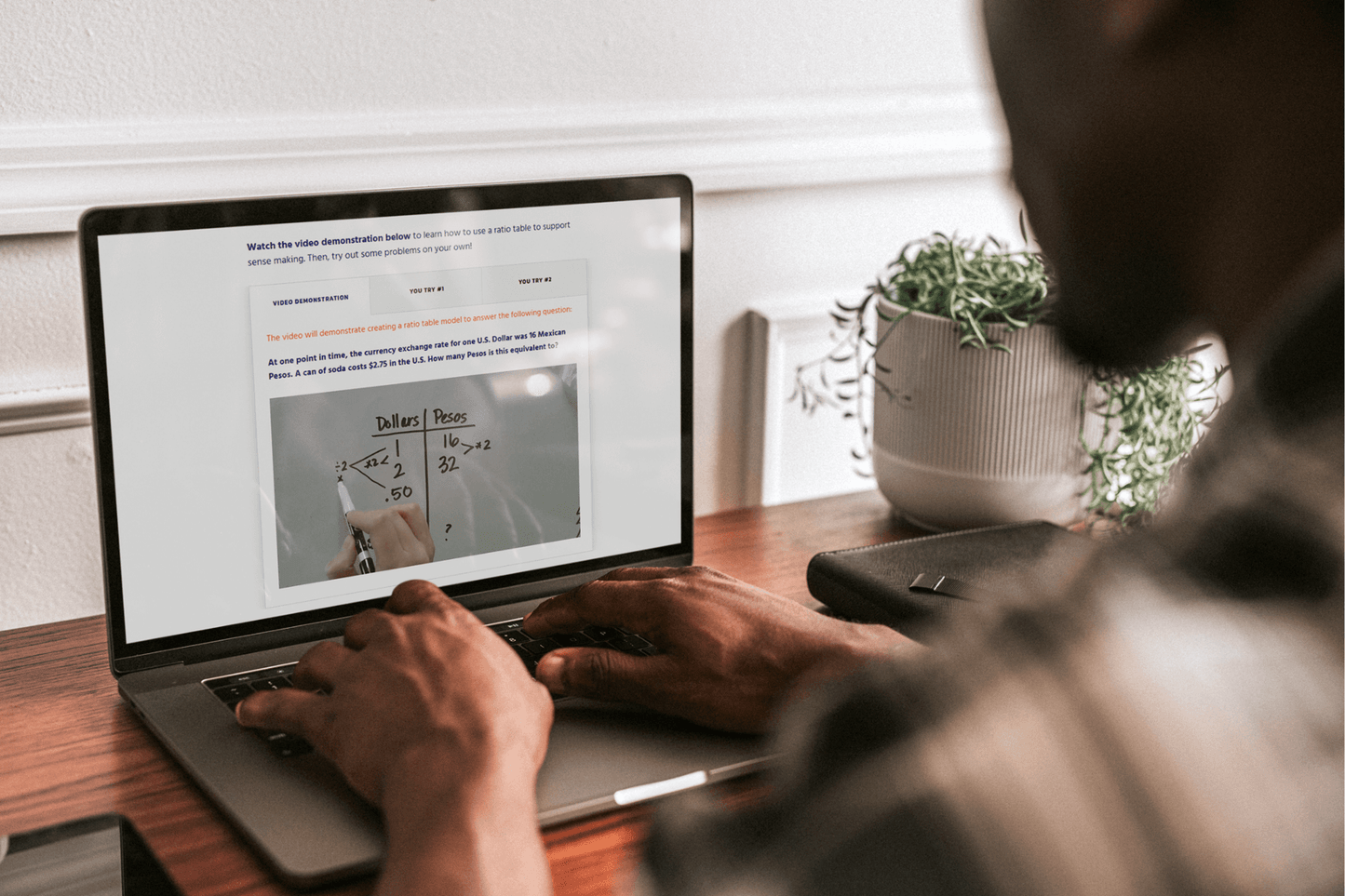 A person uses a laptop displaying a demonstration of ratio tables with a math equation related to currency conversion. A potted plant is on the wooden desk.