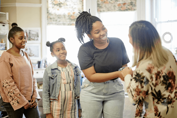 A family and an educator are smiling and shaking hands inside a classroom. Two children stand nearby, watching the warm exchange, highlighting collaboration and partnership in a supportive school setting.