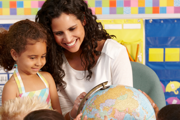 A teacher sits with a group of young children in a colorful classroom, smiling as they help them explore a globe. One child in the foreground wears a pastel striped dress and looks curiously at the globe. The background features a bright wall with colorful squares and hanging classroom organizers.