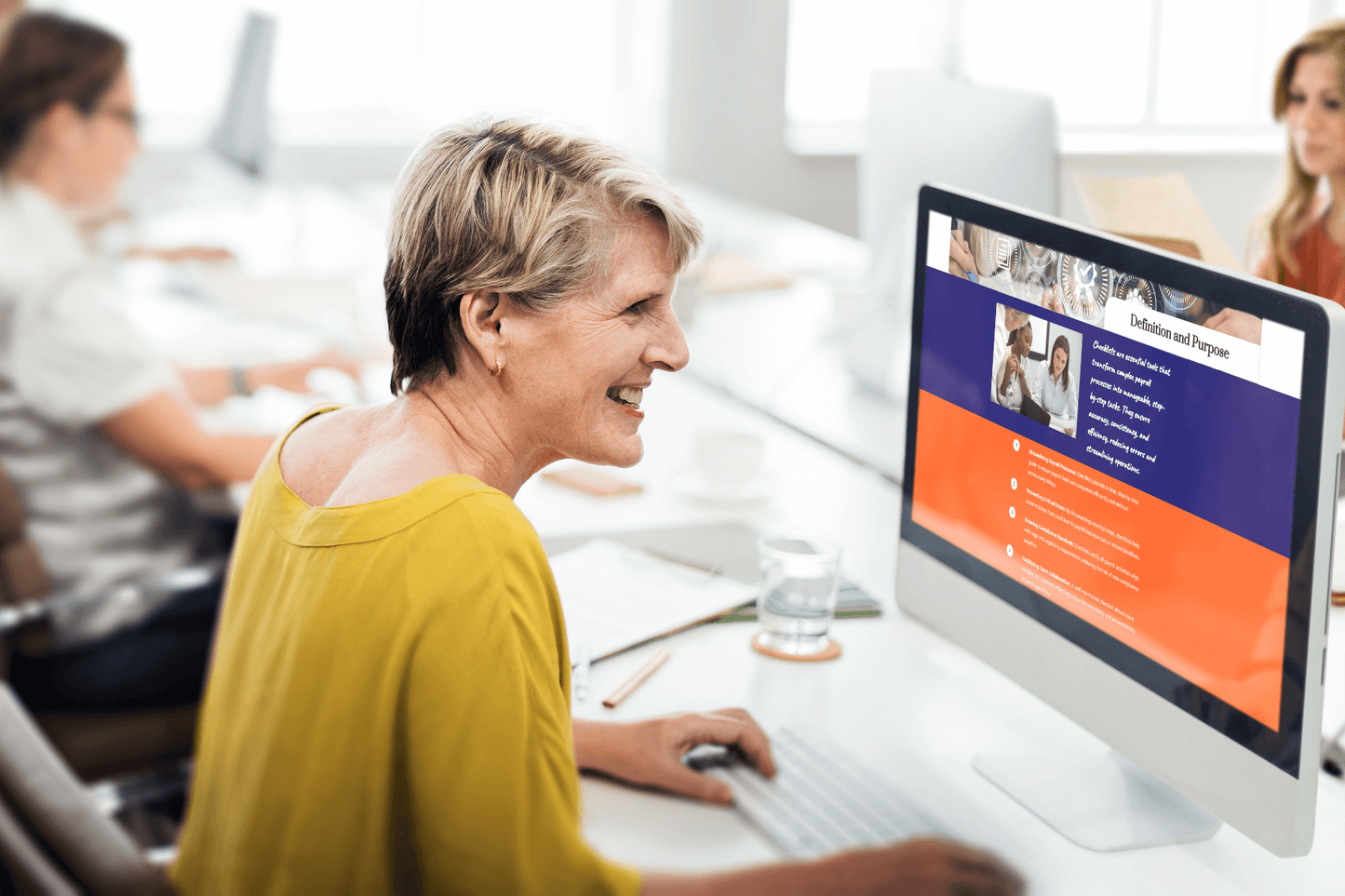 A woman in a yellow blouse smiles at a computer screen displaying a website. She's in a bright, modern office with colleagues working nearby.