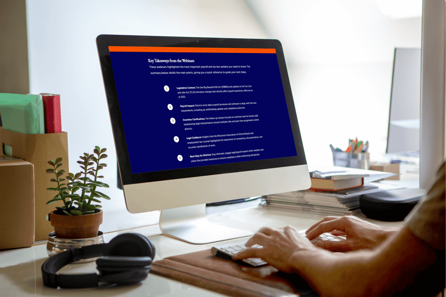 A person works at a desktop computer displaying a training module slide titled “Key Takeaways from the Webinars.” The screen shows a list of five key points related to payroll and tax law updates, covering legislation context, payroll impact, overtime clarification, legal guidance, and next steps for districts. The workspace includes a plant, headphones, and stacked books on the desk, creating an organized office environment.