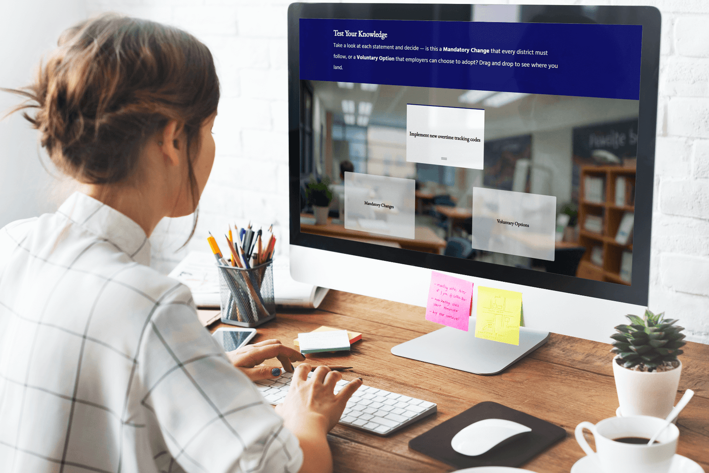 A person works at a desktop computer displaying an interactive learning activity titled “Test Your Knowledge.” The screen shows a drag-and-drop exercise asking whether a statement, such as “Implement new overtime tracking codes,” is a mandatory change or a voluntary option. On the desk are pencils in a holder, sticky notes, a smartphone, and a cup of coffee, creating a study or office environment.