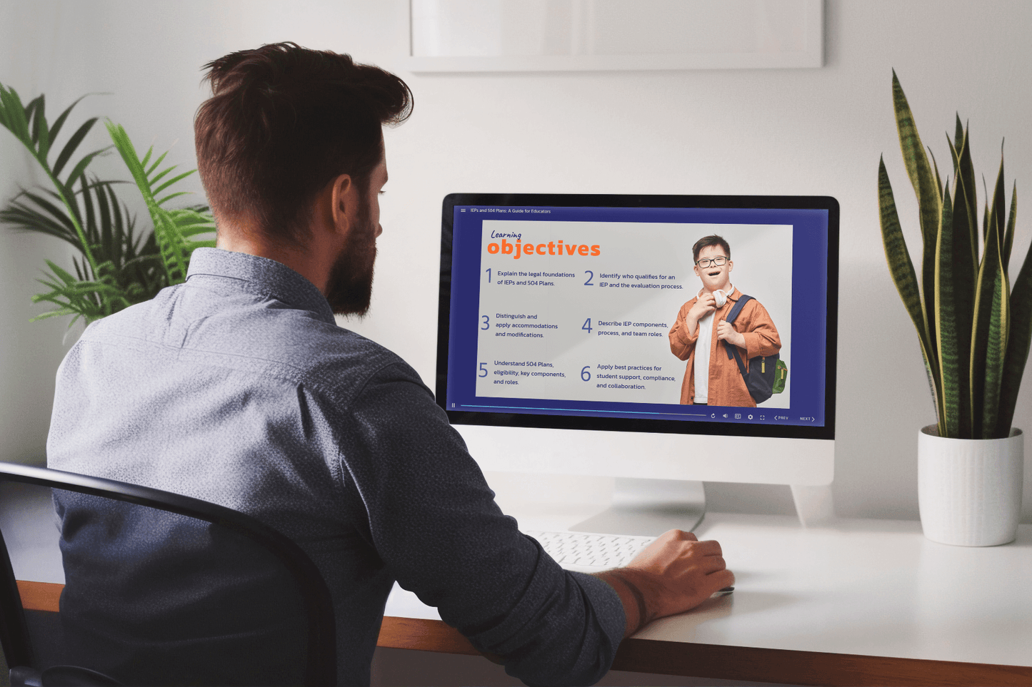 A person sits at a desk viewing an online learning module titled “IEPs and 504 Plans: A Guide for Educators” on a desktop computer. The slide on the screen is titled “Learning Objectives” and includes a list of six goals related to understanding IEPs and 504 Plans. On the right side of the slide is a photo of a smiling student wearing glasses and a backpack. The workspace features a potted plant and is brightly lit with natural light from a nearby window.