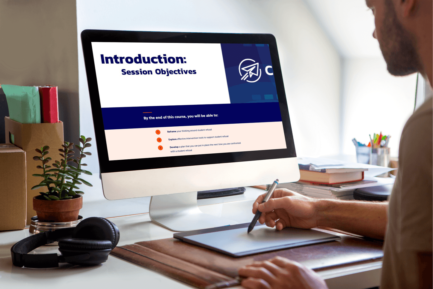 A person uses a graphics tablet at a desk. A computer screen displays a presentation titled “Introduction: Session Objectives” in a well-organized workspace.