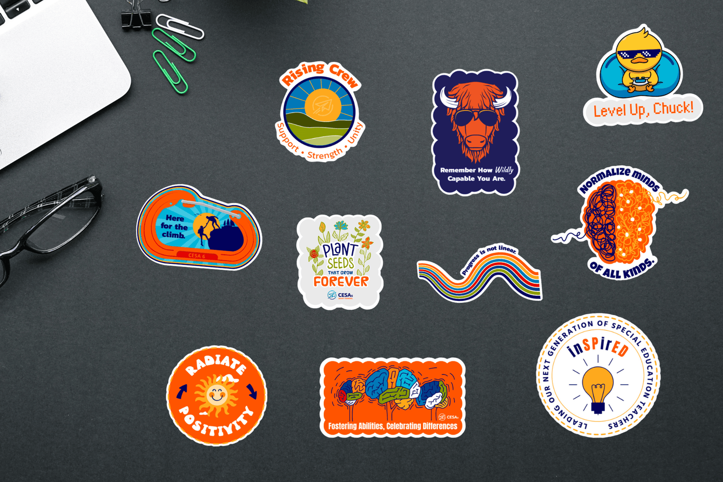 A collection of colorful stickers with motivational and inclusive messages displayed on a dark surface. The stickers include phrases like 'Radiate Positivity,' 'Normalize minds of all kinds,' 'Fostering Abilities, Celebrating Differences,' and 'Plant seeds that grow forever.' Other designs feature a buffalo with sunglasses, a duck with a gaming controller, and a sun with a smiley face. Office supplies such as paper clips and glasses are also present in the scene.