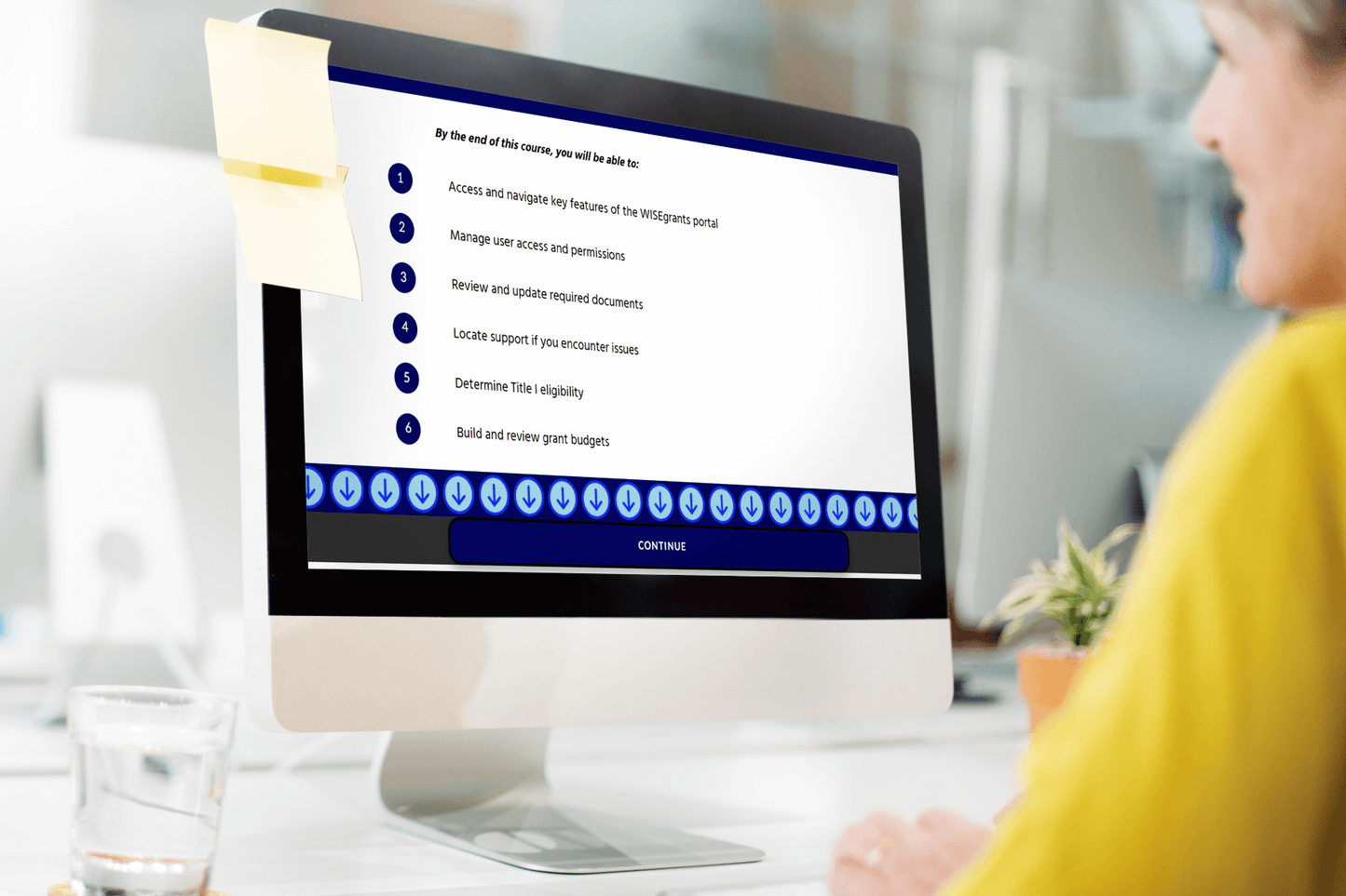 A person views a computer screen displaying an online course module with a checklist. A sticky note is attached to the screen. The scene is professional and focused.