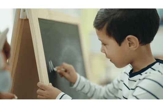 A young child is focused on writing with white chalk on a small, wooden-framed chalkboard. They are wearing a white and navy striped shirt, and his hand is steady as they forms a letter.