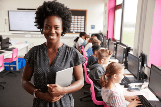 An educator stands in the foreground of a computer lab, smiling and holding a tablet. In the background, students sit at rows of computers working on assignments.
