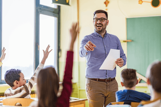 An educator standing at the front of a classroom smiles and points while holding papers. Several students seated at desks have raised hands to participate.