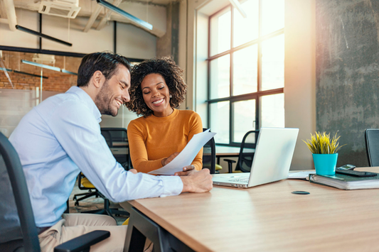 A man and woman are smiling and reviewing papers together at a desk in a bright office with a laptop and plant.