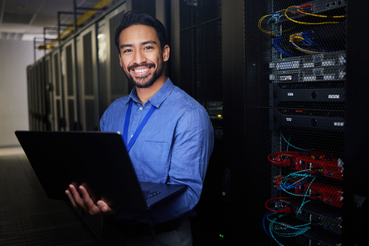 A smiling IT professional holds a laptop in a server room, surrounded by illuminated server racks and cables, wearing a blue shirt and lanyard.