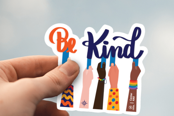 A hand holds a colorful sticker featuring the phrase "Be Kind" in bold lettering. Below, raised hands of diverse skin tones and patterns symbolize inclusivity, with one hand wearing a rainbow bracelet and others showcasing unique designs.