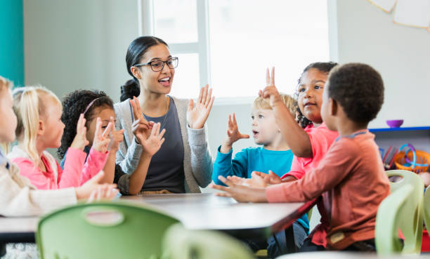 A cheerful teacher sits at a round table surrounded by young children in a brightly lit classroom. The group is engaged in a lively counting or singing activity, with the teacher and children using their fingers to show numbers.