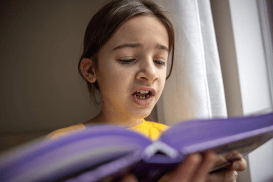 A young student is standing near a window, reading aloud from a purple book with focus and expression. The natural light highlights their face as they practice oral reading fluency. 