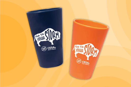 Two silicone cups are displayed against a yellow and orange gradient background. One cup is dark blue, and the other is orange. Both cups feature a white buffalo graphic with the phrase “Go to Your Storm” and the CESA 6 Smart Thinking logo beneath it.