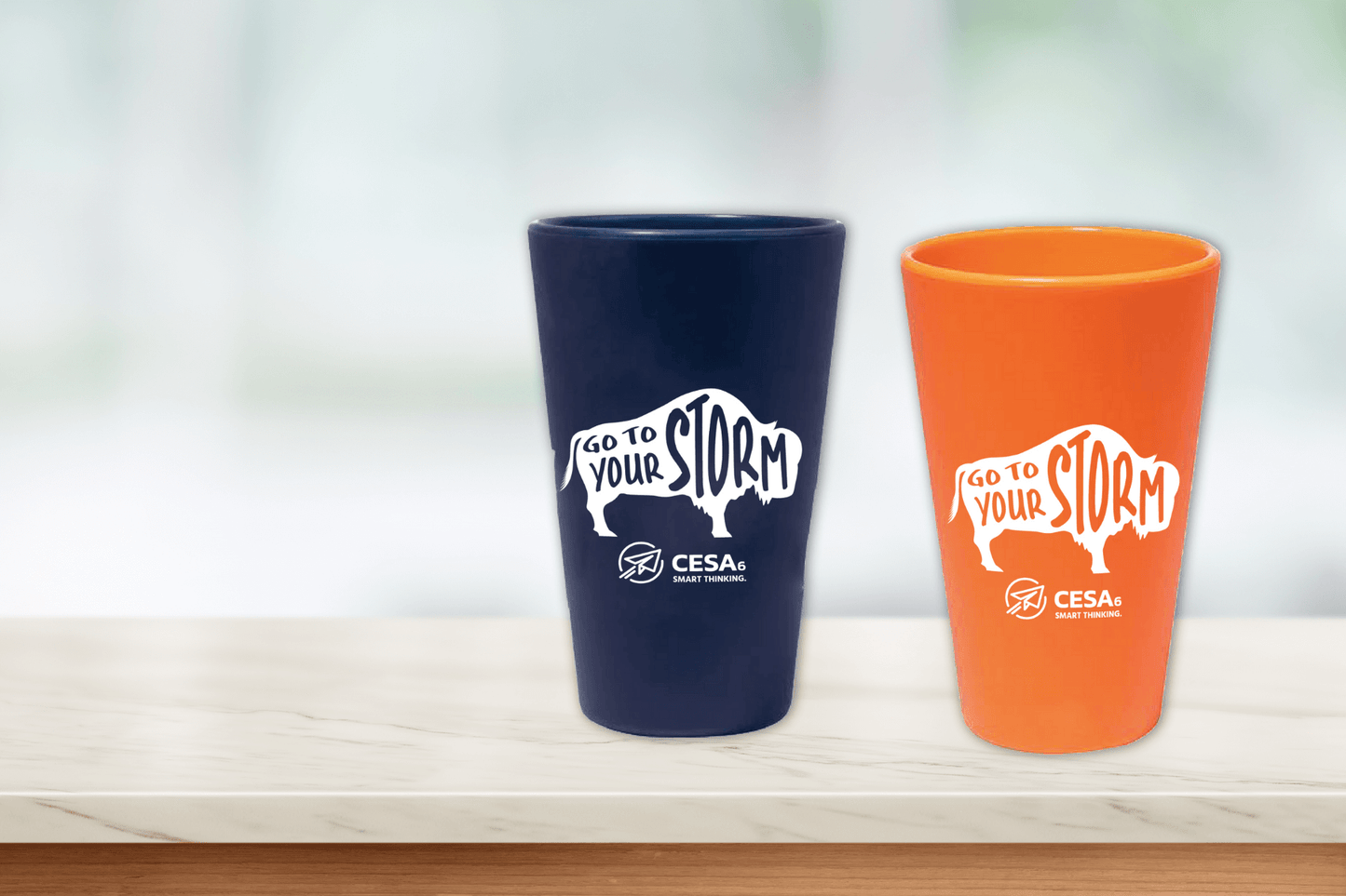 This image shows two silicone cups standing on a light-colored marble countertop with a soft, blurred background. One cup is dark blue and the other is orange. Both feature a white graphic of a buffalo with the phrase “Go to Your Storm” inside the buffalo shape. Below the buffalo, the logo for CESA 6 Smart Thinking is prominently displayed.