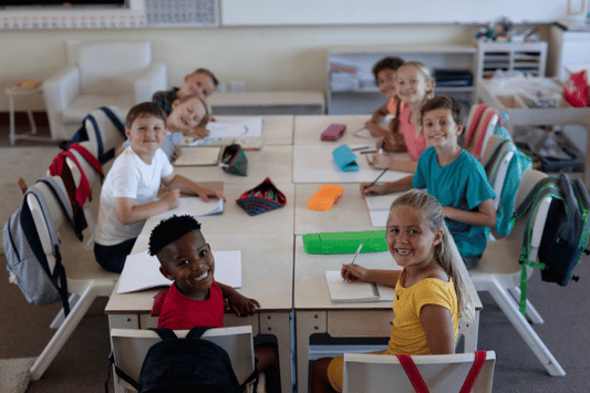 A group of young students sits around a large square arrangement of desks in a bright and organized classroom. Each child has a notebook or worksheet in front of them, and many are smiling or looking up at the camera.