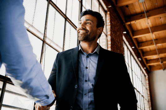 A man in a business suit smiles warmly while shaking hands with another person inside a sunlit industrial-style office with tall windows. The handshake suggests a positive professional interaction, symbolizing collaboration, agreement, or partnership.