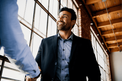 A smiling individual in a blazer and button-up shirt shakes hands with another person in a sunlit hallway with large industrial-style windows and wooden ceiling beams. The atmosphere suggests a professional or welcoming interaction, possibly a greeting or meeting.