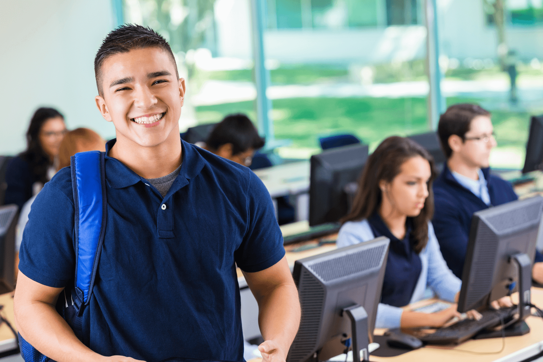 A smiling student wearing a dark blue shirt and backpack stands in a computer lab, looking at the camera. In the background, other students are seated at desktop computers, focused on their screens. The room is brightly lit with large windows showing greenery outside.