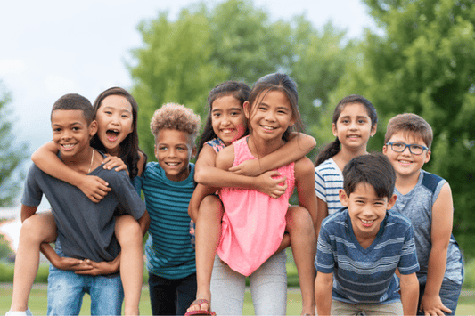 Group of smiling children standing outdoors, with two pairs giving piggyback rides, and others standing closely together in front of a blurred background of green trees.