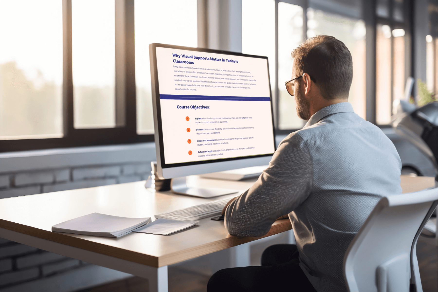 Man in an office setting working on a computer. The screen displays an educational text about visual supports in classrooms. Natural light filters through large windows.