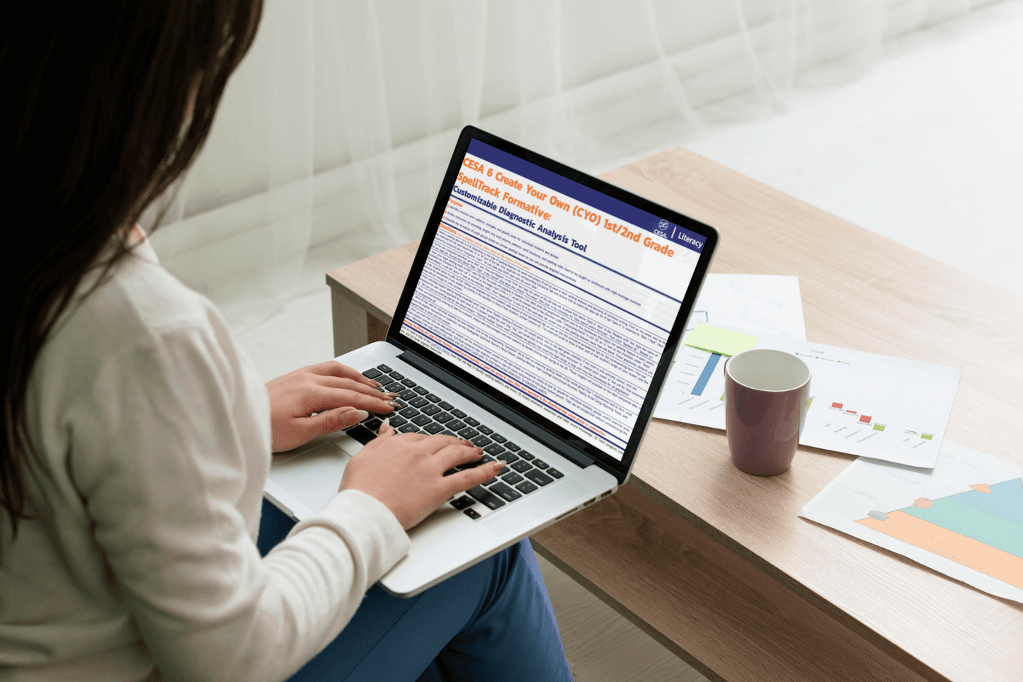 A woman uses a laptop at a light wooden table, analyzing documents with charts and a mug nearby. The scene conveys focus and productivity.