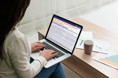 A woman uses a laptop at a light wooden table, analyzing documents with charts and a mug nearby. The scene conveys focus and productivity.
