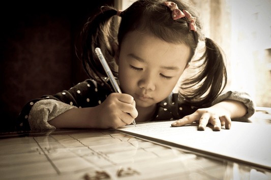 A young child with pigtails sits at a table, focused on writing in a notebook with a pen. Sunlight streams through a nearby window, illuminating the child’s concentrated expression.